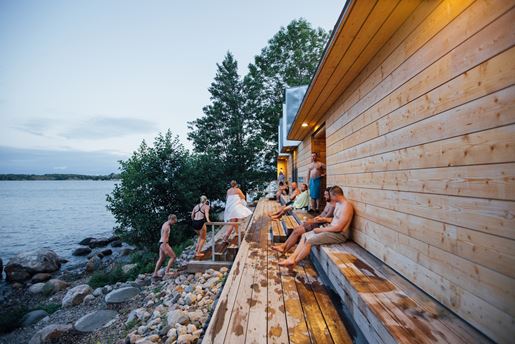 A group of people outside a sauna on the edge of a lake in Finland